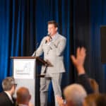 Man in a gray suit speaks at a Canine Companions podium onstage before an applauding audience