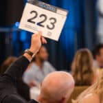 Auction bidder holding up paddle number 223 at a lively indoor event