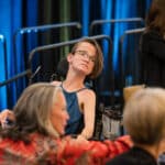 Woman in a wheelchair sits among attendees at an event, listening attentively