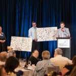 Canine Companions event with speakers onstage, a puppy vest dog, and two abstract paintings on easels