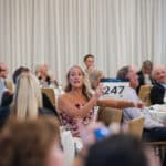 Woman bids at a charity auction in a formal dining room with seated guests and floral centerpieces