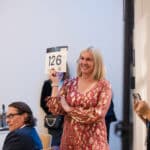 Woman smiling and holding a bid number card at an indoor event while others watch nearby