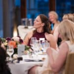 Guests seated at a formal dinner event, listening attentively around a flower-decorated table