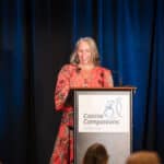 Woman in a red dress speaks at a Canine Companions podium on a stage with blue curtains
