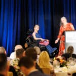 Woman in a wheelchair speaks on stage beside a podium while another woman introduces her to an audience