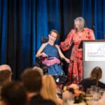 Smiling woman in a wheelchair beside a speaker on stage at a Canine Companions event