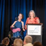 Two women at a podium during a Canine Companions event, one seated in a wheelchair, with blue stage curtains behind them
