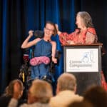 Woman in a wheelchair smiles beside a Canine Companions speaker at a podium onstage