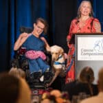 A woman in a wheelchair laughs beside a blue-vest service dog at a Canine Companions event