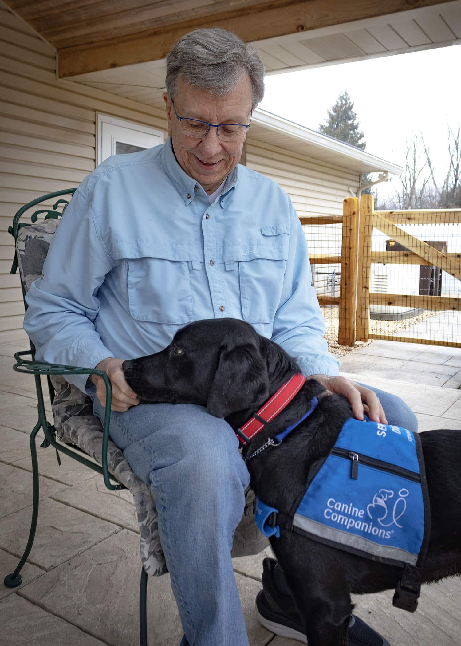 Man sitting outdoors with a black service dog in a blue Canine Companions vest resting on his lap