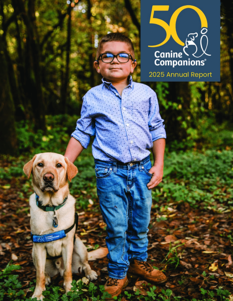 Young boy stands beside a yellow Labrador in a blue service dog vest in a wooded setting