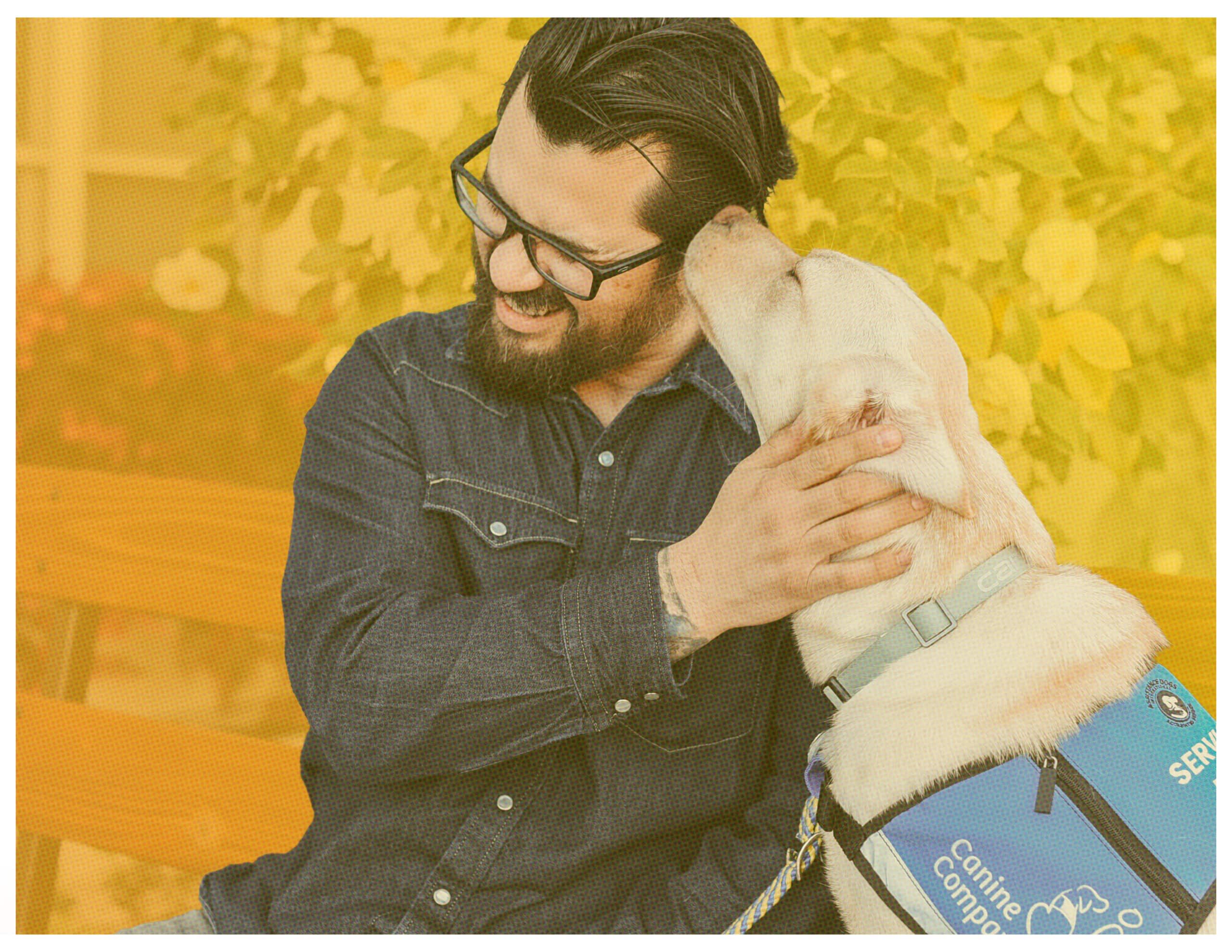 Man in glasses hugs a cream service dog wearing a blue Canine Companions vest