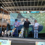 A man in a black quarter-zip sweatshirt and dark gray slacks is on a microphone while on a stage with a banner reading GOPRO behind him. He has a golden Labrador Retriever with him in a yellow Canine Companions vest. A young man with blond hair in a white shirt and blue jeans is at his left, and on his right is a woman in a lavender shirt with a black ball cap. Both women have Labrador Retrievers on leashes with them. 