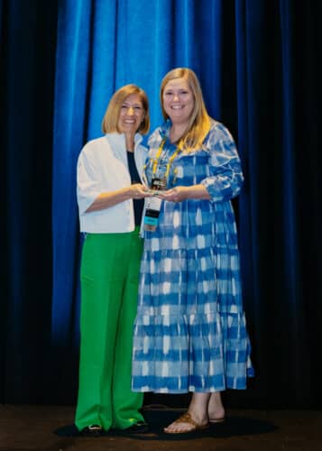 Two smiling women pose on stage while holding an award trophy in front of blue curtains