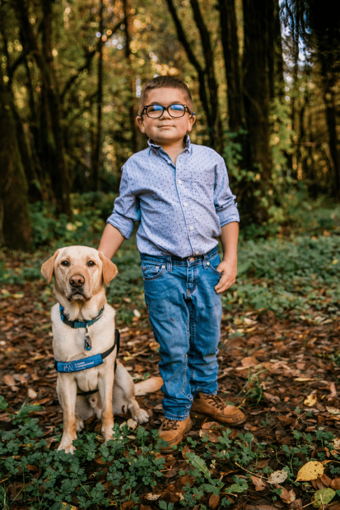 Boy standing with a yellow vest service dog in a wooded area