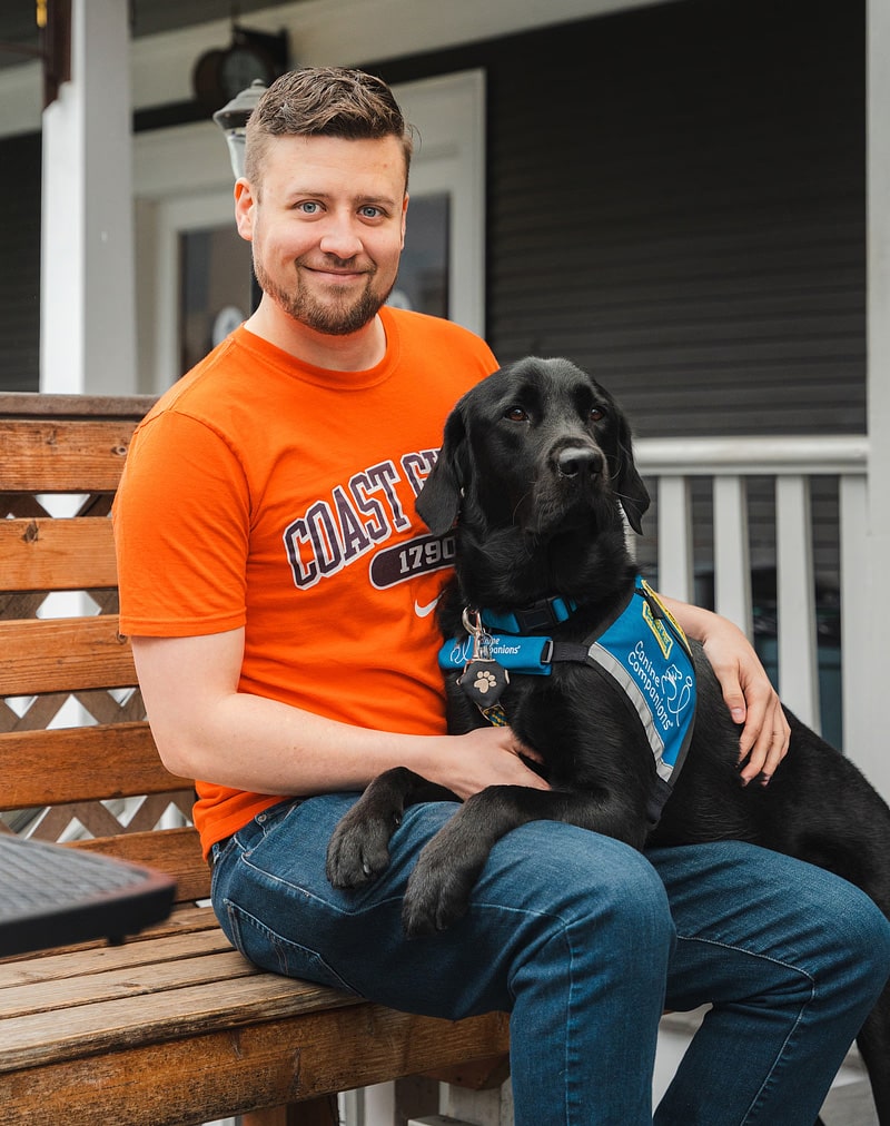 Man in an orange shirt sits with a black service dog in a blue vest on a wooden bench