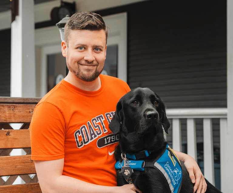 Man in an orange shirt sits with a black service dog wearing a blue Canine Companions vest