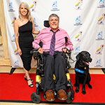 Man in a wheelchair with a black service dog in a blue vest beside him, standing with a woman on a red carpet