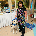 Woman smiling beside a service dog in a blue vest indoors at an event table
