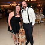 Smiling couple with a golden retriever puppy in a yellow vest at a formal indoor event
