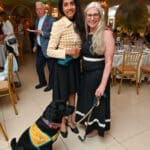 Black service dog in a yellow Canine Companions vest sits beside two smiling women at an indoor event