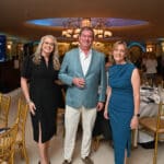 Three adults pose at a formal indoor event under ornate chandeliers