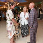 A service dog in a blue vest sits between three people chatting at an elegant indoor event