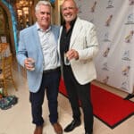 Two men in blazers smile and hold drinks at a Canine Companions event with a red carpet and logo backdrop