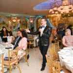 Man in a sequined tuxedo jacket speaks into a microphone at a Canine Companions banquet with seated guests