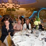 Guests seated at a formal gala in a chandelier-lit dining room, with a small dog under a table in the background