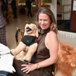 Woman smiling while holding a plush dog at a formal indoor event with a horse-costumed figure nearby