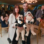 Smiling couple holding two plush Canine Companions dogs in formal outfits at a banquet event