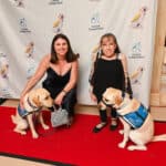 Two women pose on a red carpet with two Canine Companions service dogs in blue vests