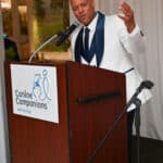 Man in a white tuxedo speaking at a Canine Companions podium with a microphone
