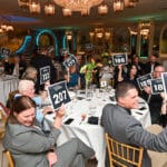 Guests at a fundraising banquet hold up bidding signs in a chandelier-lit ballroom