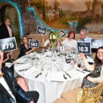 Guests at a gala table hold auction paddle signs in front of a painted mural backdrop