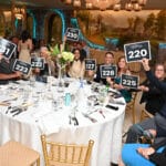 Guests at a gala table hold numbered auction paddles and smile for the camera