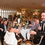 Man in sequined jacket speaking into a microphone at a banquet table of guests, with a small dog partly visible below