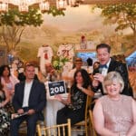 Auction host speaking into microphone at a formal Canine Companions event with seated guests and sports memorabilia backdrop