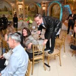 A man in a sequined jacket leans over a table, speaking into a microphone at a formal Canine Companions event