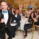 Man in a sequined tuxedo jacket speaks into a microphone at a lively banquet with smiling guests at round tables