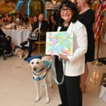 Service dog in a blue vest stands beside a smiling woman at an indoor event, with guests seated behind them