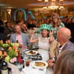 Guests at a banquet with floral table settings, including a woman trying on a Statue of Liberty crown