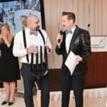 Two men in formalwear chat at a Canine Companions event, one holding a giant fork, with a podium in the background