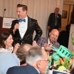 Man in a black sequined jacket speaks into a microphone at a Canine Companions event with seated guests and flowers