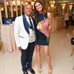 Man in a white tuxedo and woman in a navy dress smile together at an elegant indoor event, with the woman holding a bouquet