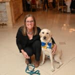 Woman crouches beside a yellow-vested service dog in a formal banquet hall