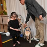 A man in a blue service-dog vest gently pets a yellow Labrador beside a woman holding a phone