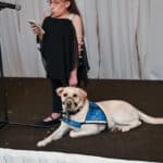 Woman speaking at a microphone while a blue-vested service dog lies beside her on stage
