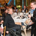 Service dog in a blue vest greets a man speaking into a microphone at a formal event